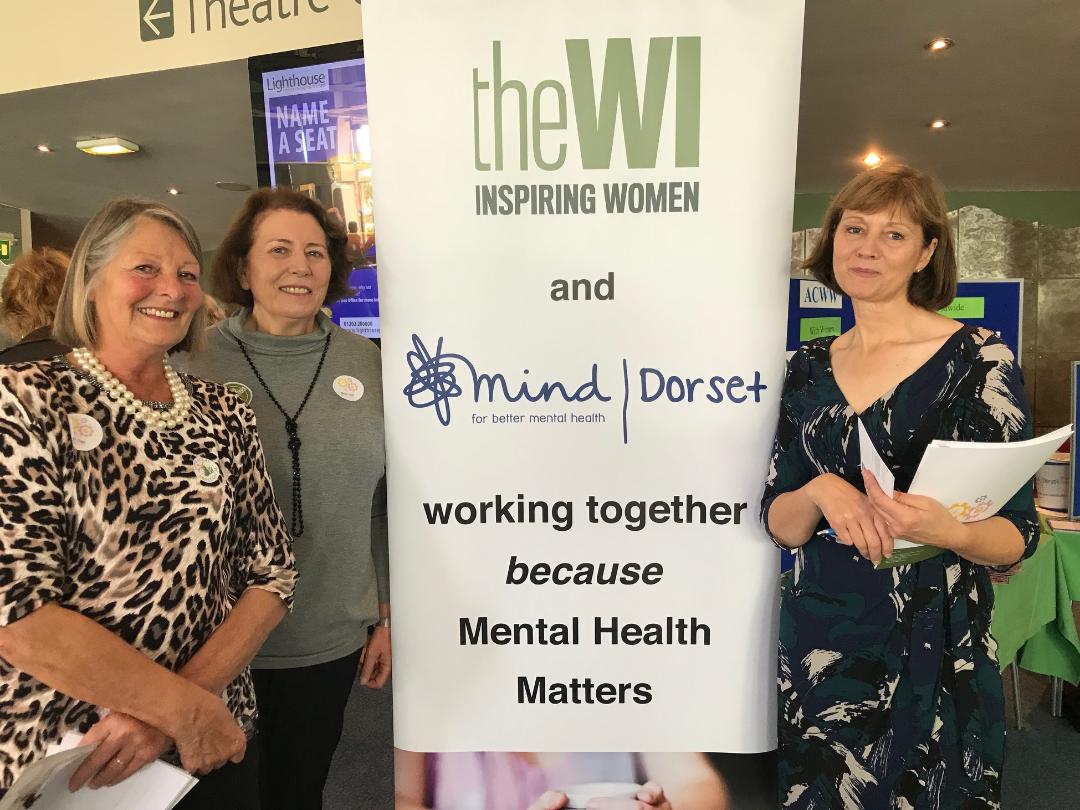 Three women standing next to a banner that reads "The WI and mind | Dorset"